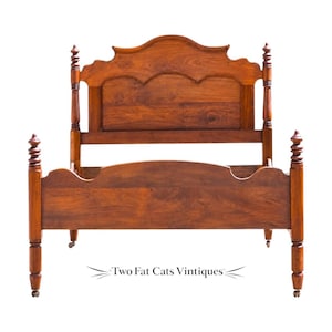 May include: A brown wooden bed frame featuring a tall headboard with a curved top edge and a matching lower footboard. The frame includes four vertical posts with decorative turned finials and small wheels at the base. Text reads Two Fat Cats Vintiques.