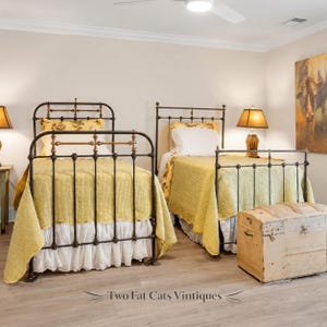 May include: Two twin-sized metal bed frames with dark brown and brass-colored accents, each dressed in yellow quilted coverlets, white ruffled bed skirts, and white pillows. A light-colored wooden trunk sits at the foot of one bed.
