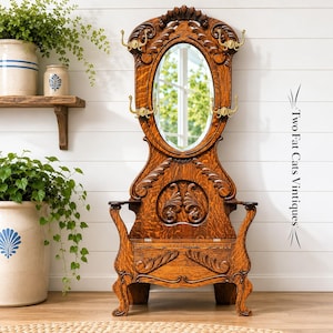 May include: A tall, dark wood hall tree featuring an oval mirror, intricate carved leaf details, and four brass hooks. The base includes a storage compartment with a hinged lid. The text Two Fat Cats Vintiques is visible on the right side.