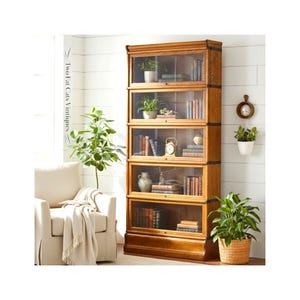 May include: A tall, five-tier wooden bookcase with a medium brown finish, featuring individual glass-front doors for each shelf. The unit is constructed with dark metal hardware accents and contains various books, small potted plants, and a clock.