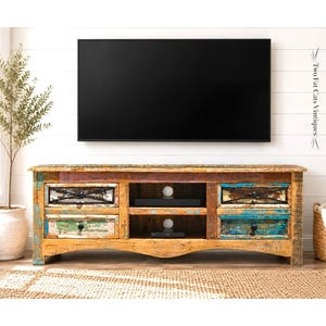 May include: A rectangular wooden media console featuring a distressed, multi-colored finish with patches of blue, green, and natural wood tones. It includes four drawers with black metal hardware and two open central shelves. Text reads Two Fat Cats Vintiques.