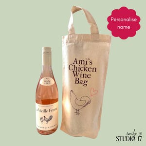 May include: A bottle of rosé wine and a canvas wine bag. The wine bottle has a light pink colour and a label. The wine bag has a handle and the text "Ami's Chicken Wine Bag" with a drawing of a chicken. The bag is made of natural canvas.
