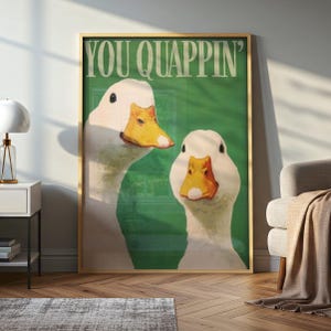 May include: A framed art print featuring two white ducks with orange beaks against a green background. The text "YOU QUAPPIN'" is displayed at the top. The artwork is in a gold frame, adding a touch of elegance.