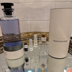 May include: A collection of perfume bottles and vials, including a large bottle of M&eacute;t&eacute;ore by Louis Vuitton. The bottles vary in size, with labels indicating 1 ml, 3 ml, 5 ml, and 15 ml. The packaging includes a cylindrical box.