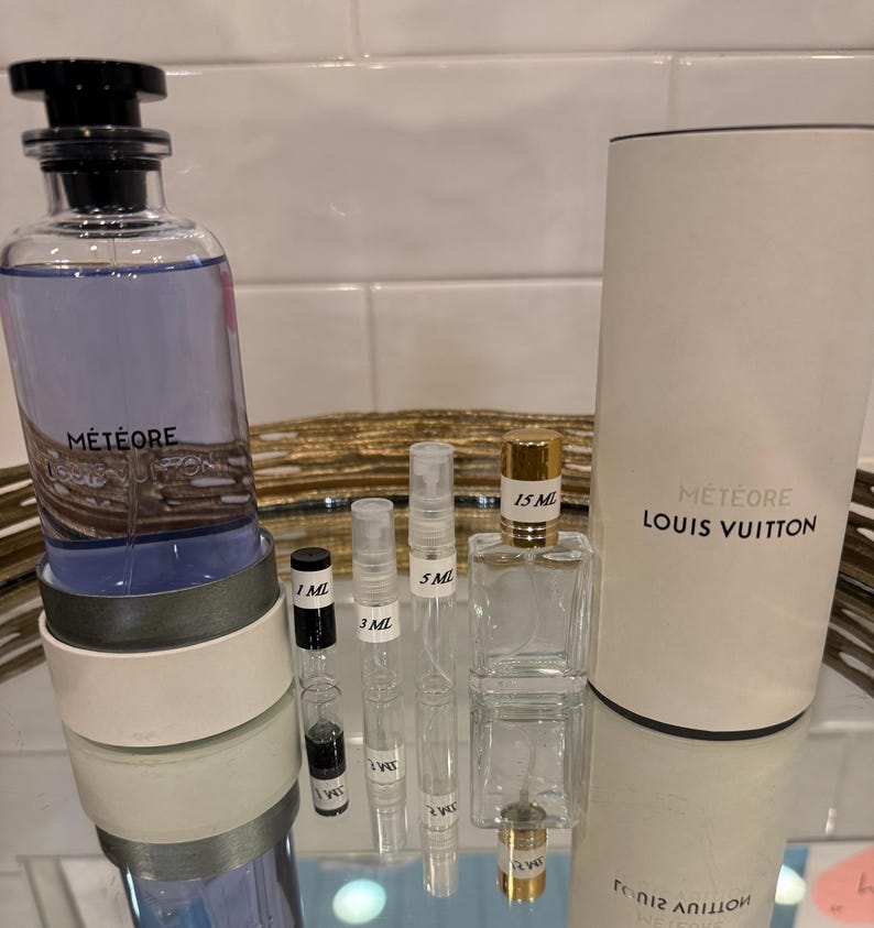 May include: A collection of perfume bottles and vials, including a large bottle of M&eacute;t&eacute;ore by Louis Vuitton. The bottles vary in size, with labels indicating 1 ml, 3 ml, 5 ml, and 15 ml. The packaging includes a cylindrical box.