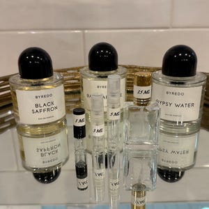 May include: An assortment of Byredo perfume bottles and samples. The image features three full-size bottles with black caps and white labels, alongside several small sample vials. The bottles are labeled "Black Saffron" and "Gypsy Water".