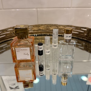 May include: An assortment of perfume bottles and vials on a reflective surface. The bottles vary in size and shape, including a square orange bottle with gold accents and the text "Maison Francis Kurkdjian Paris". Several small vials are labeled with measurements like 1 ML, 3 ML, and 5 ML.