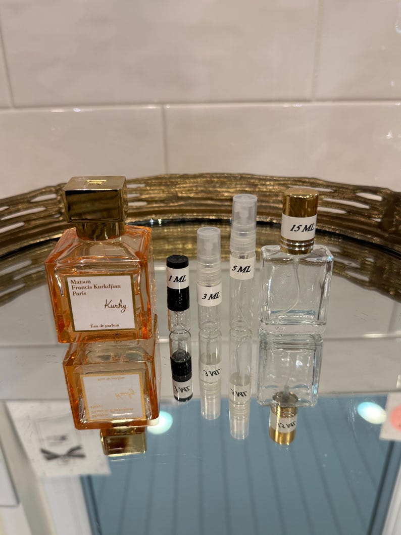 May include: An assortment of perfume bottles and vials on a reflective surface. The bottles vary in size and shape, including a square orange bottle with gold accents and the text "Maison Francis Kurkdjian Paris". Several small vials are labeled with measurements like 1 ML, 3 ML, and 5 ML.