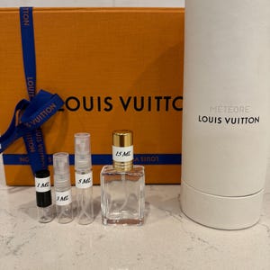 May include: An orange Louis Vuitton box with a blue ribbon, and several perfume bottles. The bottles include sizes 1 ml, 3 ml, 5 ml, and 15 ml. A white cylindrical container with "M&eacute;t&eacute;ore Louis Vuitton" is also present.