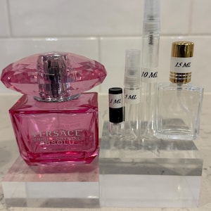 May include: A collection of perfume bottles and vials. A large, pink, faceted bottle with a silver cap is next to several clear vials labeled 1 mL, 5 mL, 10 mL, and 15 mL. The large bottle has the text "Versace Bright Crystal Absolu".