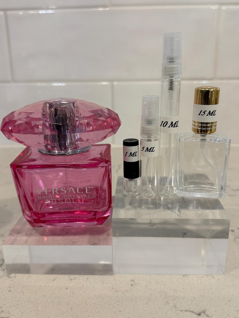 May include: A collection of perfume bottles and vials. A large, pink, faceted bottle with a silver cap is next to several clear vials labeled 1 mL, 5 mL, 10 mL, and 15 mL. The large bottle has the text "Versace Bright Crystal Absolu".