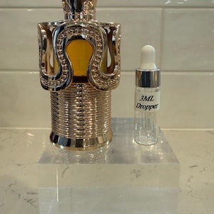 May include: A decorative perfume bottle with a gold-colored, ornate design. The bottle has a unique shape and is accompanied by a small, clear 3mL dropper bottle. The perfume bottle sits on a clear acrylic block.
