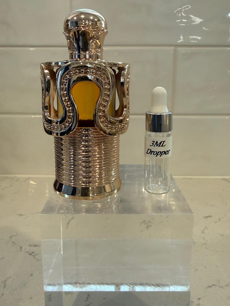 May include: A decorative perfume bottle with a gold-colored, ornate design. The bottle has a unique shape and is accompanied by a small, clear 3mL dropper bottle. The perfume bottle sits on a clear acrylic block.