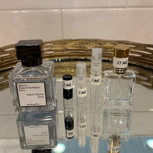 May include: An assortment of perfume bottles and vials on a reflective surface. The bottles include a square glass bottle with a black cap and the text "Aqua Celestia forte". There are also several small vials labeled 1 ML, 3 ML, 5 ML, and 15 ML.
