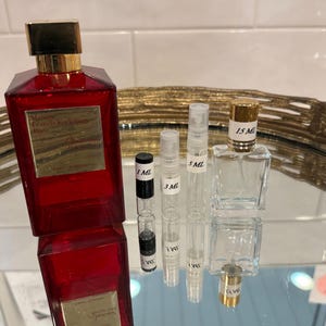 May include: A collection of perfume bottles and vials. A large, red rectangular perfume bottle with a gold cap is next to several smaller, clear vials labeled 1 ML, 3 ML, 5 ML, and 15 ML. The bottles are arranged on a reflective surface.