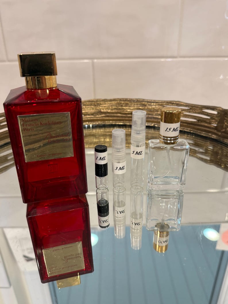 May include: A collection of perfume bottles and vials. A large, red rectangular perfume bottle with a gold cap is next to several smaller, clear vials labeled 1 ML, 3 ML, 5 ML, and 15 ML. The bottles are arranged on a reflective surface.