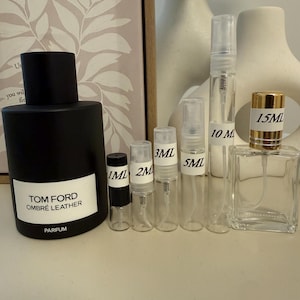 May include: A large black Tom Ford Ombré Leather parfum bottle alongside a row of five clear glass spray sample vials labeled 1ml, 2ml, 3ml, 5ml, and 10ml, and a square 15ml glass perfume bottle with a gold cap, all arranged on a flat surface.