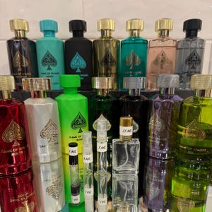May include: Collection of perfume bottles in various colors, including black, green, and red, with gold and silver accents. Each bottle features a spade symbol and the text "GAME OF SPADES". Several sample vials are also visible.