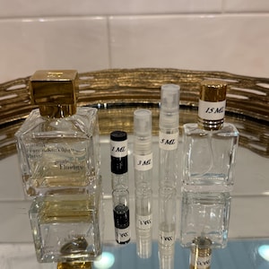 May include: An assortment of perfume bottles and vials. Two square glass bottles with gold caps are labeled "gentle Fluidity". Several clear vials are labeled with measurements: 1 ML, 3 ML, 5 ML, and 15 ML. The bottles and vials are arranged on a reflective surface.