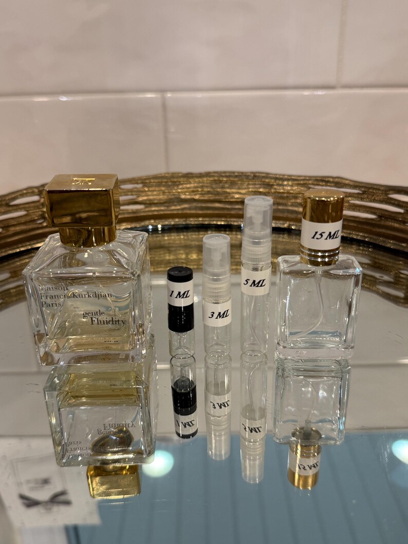 May include: An assortment of perfume bottles and vials. Two square glass bottles with gold caps are labeled "gentle Fluidity". Several clear vials are labeled with measurements: 1 ML, 3 ML, 5 ML, and 15 ML. The bottles and vials are arranged on a reflective surface.