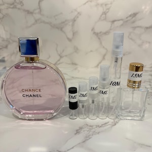 Puede incluir: Una colecci&oacute;n de frascos y atomizadores de perfume. Un frasco de perfume grande, redondo y rosa con una tapa azul y plateada est&aacute; etiquetado como "Chance Chanel". Varios viales peque&ntilde;os y transparentes est&aacute;n etiquetados con medidas de 1 ml a 15 ml.