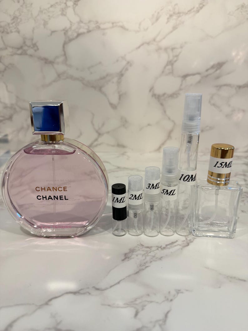 Puede incluir: Una colecci&oacute;n de frascos y atomizadores de perfume. Un frasco de perfume grande, redondo y rosa con una tapa azul y plateada est&aacute; etiquetado como "Chance Chanel". Varios viales peque&ntilde;os y transparentes est&aacute;n etiquetados con medidas de 1 ml a 15 ml.