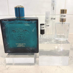 May include: A teal Versace Eros Parfum bottle with a blue cap and a Medusa head emblem, alongside several clear perfume sample vials of 1 mL, 5 mL, 10 mL, and 15 mL. The bottles are displayed on clear acrylic blocks.