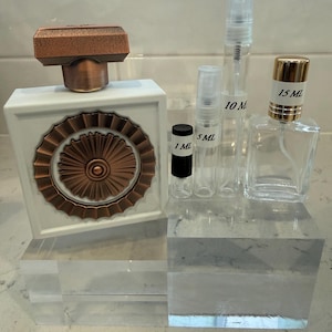 May include: A white perfume bottle with a copper-coloured top and decorative front, alongside several clear glass spray bottles labelled 1 ml, 5 ml, 10 ml, and 15 ml. The bottles are displayed on clear acrylic blocks.