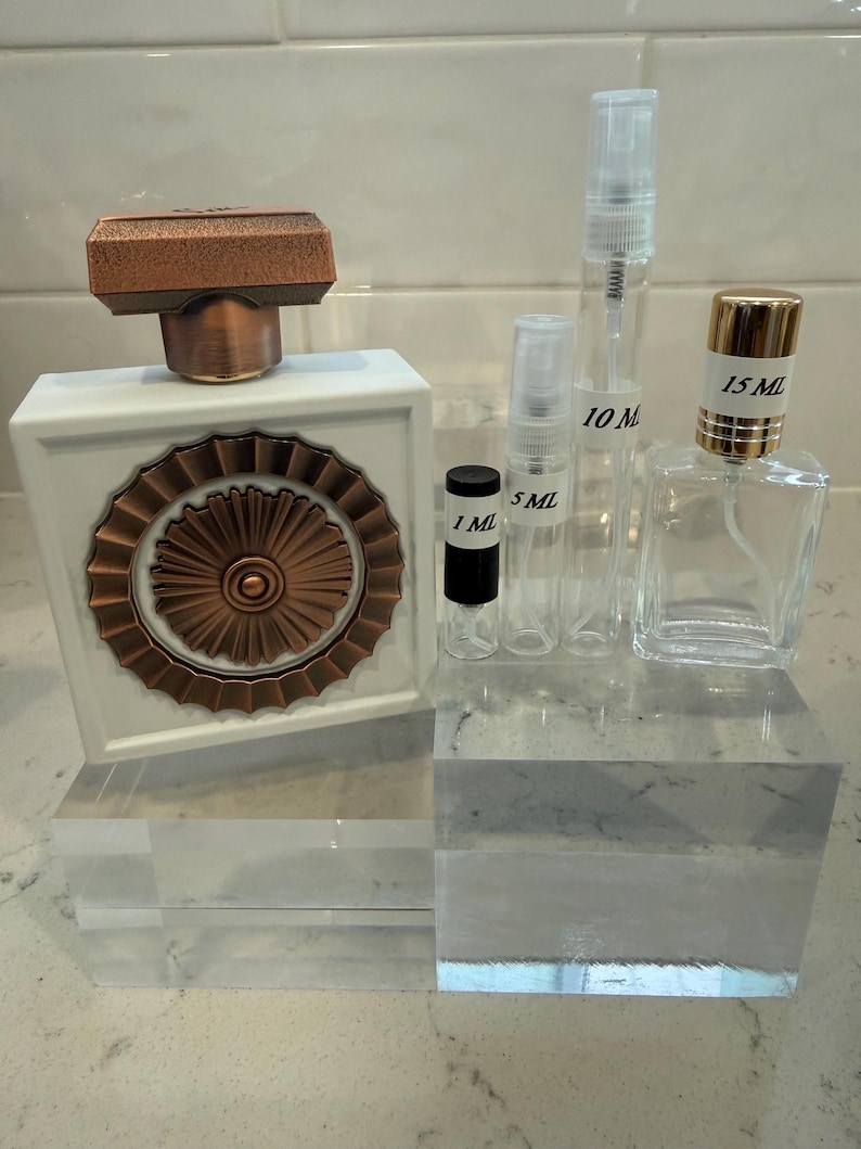 May include: A white perfume bottle with a copper-coloured top and decorative front, alongside several clear glass spray bottles labelled 1 ml, 5 ml, 10 ml, and 15 ml. The bottles are displayed on clear acrylic blocks.