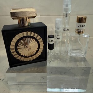 May include: A black perfume bottle with a gold cap and a gold sunburst design, alongside several clear glass spray bottles labeled 1 ml, 5 ml, 10 ml, and 15 ml. The bottles are displayed on clear acrylic blocks.