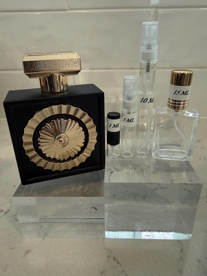 May include: A black perfume bottle with a gold cap and a gold sunburst design, alongside several clear glass spray bottles labeled 1 ml, 5 ml, 10 ml, and 15 ml. The bottles are displayed on clear acrylic blocks.