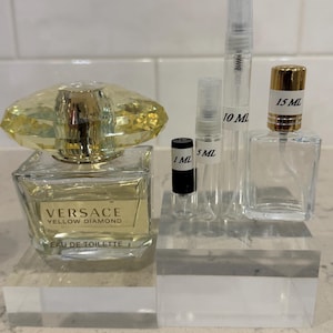 May include: A collection of perfume bottles and vials. A square bottle of Versace Yellow Diamond Eau de Toilette sits next to several clear vials labeled 1 mL, 5 mL, 10 mL, and 15 mL. The Versace bottle has a yellow crystal-like top.