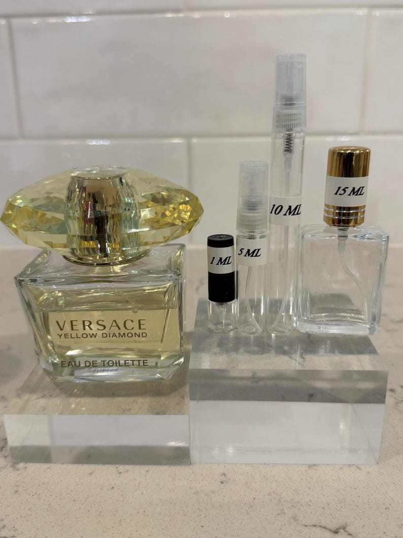 May include: A collection of perfume bottles and vials. A square bottle of Versace Yellow Diamond Eau de Toilette sits next to several clear vials labeled 1 mL, 5 mL, 10 mL, and 15 mL. The Versace bottle has a yellow crystal-like top.