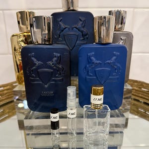 May include: An assortment of perfume bottles and vials. Several dark blue bottles with silver caps and a horse emblem are displayed. Smaller clear vials with black and gold labels are also present, with sizes ranging from 1 mL to 15 mL.