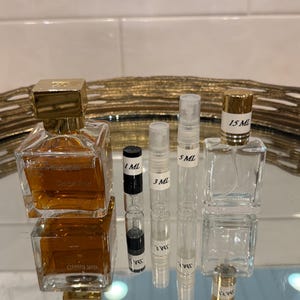 May include: An assortment of perfume bottles and vials. The image features a large, square perfume bottle with a gold cap, alongside smaller vials labeled 1 mL, 3 mL, 5 mL, and a 15 mL bottle. The bottles are filled with a light amber liquid.