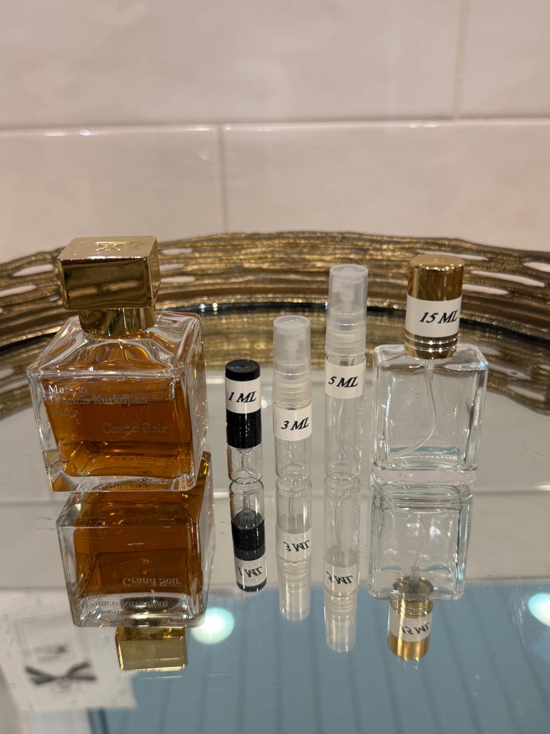 May include: An assortment of perfume bottles and vials. The image features a large, square perfume bottle with a gold cap, alongside smaller vials labeled 1 mL, 3 mL, 5 mL, and a 15 mL bottle. The bottles are filled with a light amber liquid.