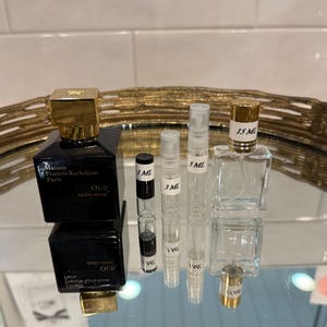 May include: A collection of perfume bottles and vials. A black square perfume bottle with a gold cap is next to several clear vials labeled 1 ML, 3 ML, 5 ML, and a larger 15 ML bottle. The bottles are on a reflective surface.