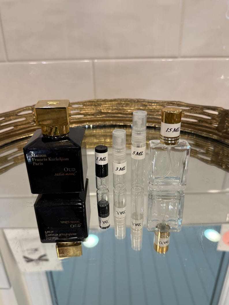 May include: A collection of perfume bottles and vials. A black square perfume bottle with a gold cap is next to several clear vials labeled 1 ML, 3 ML, 5 ML, and a larger 15 ML bottle. The bottles are on a reflective surface.