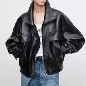 Könnte beinhalten: Eine schwarze Lederjacke mit breitem Kragen, langen Ärmeln und zwei aufgesetzten Pattentaschen vorne. Die Jacke hat einen silbernen Reißverschluss, Metalldruckknöpfe am Kragen und Saum und wird über einem weißen Tanktop und blauen Jeans getragen.