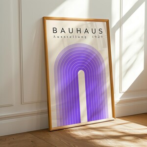 Op de afbeelding: Een ingelijste kunstdruk met een paars boogontwerp. De print heeft de tekst "BAUHAUS Ausstellung 1923" in het zwart. Het kunstwerk zit in een houten lijst en leunt tegen een witte muur en een houten vloer.