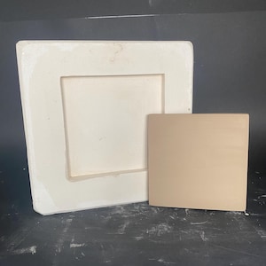 May include: A square, off-white ceramic mold with a recessed square interior, next to a smaller, tan square tile. The mold has a rough, textured surface. The tile is smooth and appears to be made of ceramic or a similar material. The items are on a dark surface.