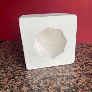 Könnte beinhalten: Eine weiße, geometrisch geformte Form mit einer facettierten, achteckigen Öffnung. Die Form hat eine Würfelform und steht auf einer gesprenkelten, braunen Oberfläche mit rotem Hintergrund.