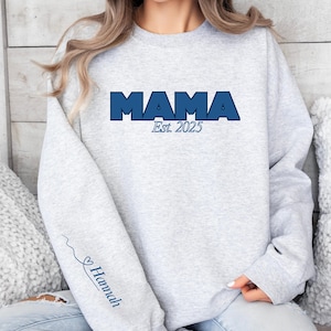 May include: A light gray sweatshirt with the word "MAMA" in bold, dark blue letters across the chest, with "Est. 2025" below. The left sleeve has a blue heart and the name "Hannah" in script.