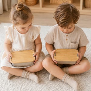 Peut inclure: Deux bo&icirc;tes &agrave; d&eacute;jeuner rectangulaires avec des couvercles en bambou clair et des bases en m&eacute;tal argent&eacute;. Chaque bo&icirc;te comporte deux petits loquets m&eacute;talliques lat&eacute;raux pour fixer le couvercle. Elles sont tenues par deux enfants assis.
