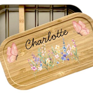 Peut inclure: Une bo&icirc;te &agrave; d&eacute;jeuner rectangulaire en acier inoxydable avec un couvercle en bambou amovible. Le couvercle porte le pr&eacute;nom Charlotte en &eacute;criture noire, entour&eacute; d'une illustration &agrave; l'aquarelle de papillons roses et de fleurs sauvages.