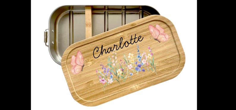 Peut inclure: Une bo&icirc;te &agrave; d&eacute;jeuner rectangulaire en acier inoxydable avec un couvercle en bambou amovible. Le couvercle porte le pr&eacute;nom Charlotte en &eacute;criture noire, entour&eacute; d'une illustration &agrave; l'aquarelle de papillons roses et de fleurs sauvages.