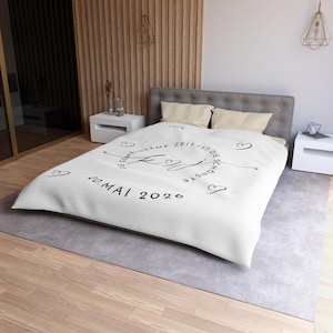 May include: White bedspread with black script and heart designs. The text reads "Die gemeinsame Zeit ist die schönste" and "20. Mai 2020". The bedspread is on a bed with gray headboard and pillows.
