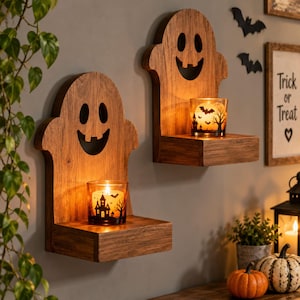 May include: Two wall-mounted wooden shelves shaped like ghosts with cut-out eyes and mouths. Each shelf holds a glass candle holder featuring a black silhouette of a haunted house, trees, and bats, with a glowing candle inside the glass.