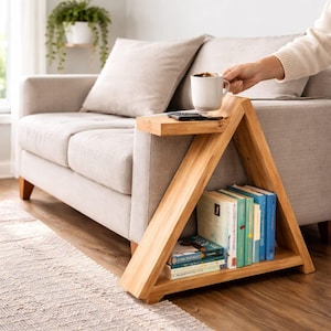 May include: A wooden side table with a unique A-frame design. The table is light brown and has a small shelf on top with a white mug and remote. The lower section holds several books. The table is next to a beige sofa.