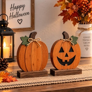 May include: Two wooden pumpkin decorations standing on rectangular bases. One pumpkin is plain orange with a green leaf and twine bow, while the other features a black jack-o'-lantern face cutout, a green leaf, and a twine bow on the stem.